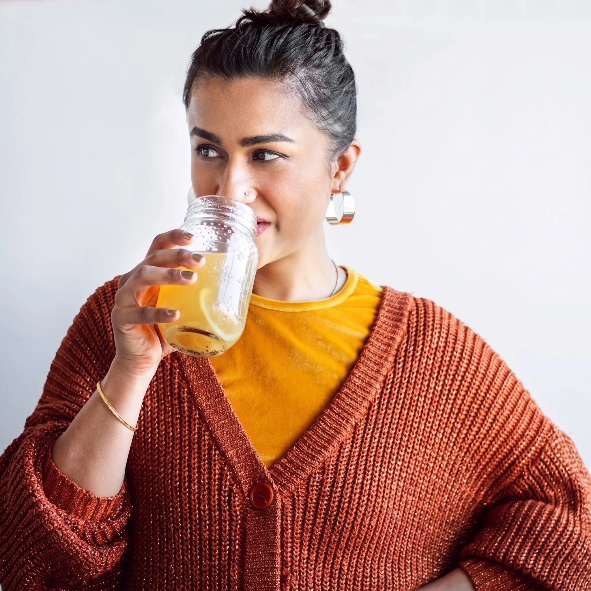 Mithalee Rawat, founder of Shorba Broth Bar, sipping broth from a glass jar in an orange knit cardigan